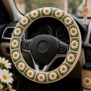 May include: A steering wheel cover featuring a repeating crochet pattern of white daisy motifs with yellow centers on a sage green square background, bordered by cream-colored yarn. The cover is fitted onto a black car steering wheel.