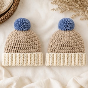 May include: Two matching beanies featuring a tan crocheted crown and a cream-colored ribbed brim. Each hat is topped with a textured, round, periwinkle blue pom-pom. The hats are constructed with a visible yarn stitch pattern throughout the crown.