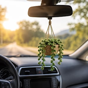 May include: A crocheted hanging plant ornament featuring a tan basket and green trailing vines with small round leaves. The plant is suspended by tan yarn strings from a central knot, designed to hang from a vehicle rearview mirror.