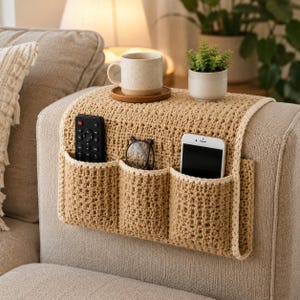 May include: A beige crochet sofa armrest organizer featuring three deep pockets draped over the side of a couch. The organizer holds a black remote control, a pair of dark-rimmed glasses, and a white smartphone. A ceramic mug and plant sit on top.