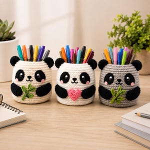 Peut inclure: Trois porte-crayons cylindriques en crochet en forme de pandas. Deux sont de couleur crème avec des oreilles, des yeux et des bras noirs, tenant des brins de bambou crochetés verts. Le centre est blanc avec un cœur rose et des traits de panda noirs.
