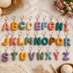 May include: A collection of twenty-six individual crochet letter keychains, each featuring a silver metal ring. The letters A through Z are crafted in various solid colours including pink, yellow, green, cream, purple, blue, and orange, arranged in three rows.