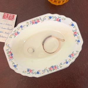 May include: An oval ceramic dish with a cream-colored center and a scalloped edge featuring a floral pattern of pink roses and small blue flowers. Inside the dish rests a thin silver-toned ring and a rose gold-toned hinged bracelet with a chain.