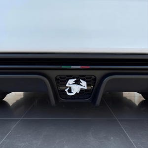 Könnte beinhalten: Ein schwarzer Heckstoßstangendiffusor mit einem zentralen Wabengitter, in dessen Mitte ein weißes Skorpion-Emblem angebracht ist. Eine schmale horizontale Zierleiste über dem Gitter zeigt einen kleinen Streifen in den italienischen Farben.