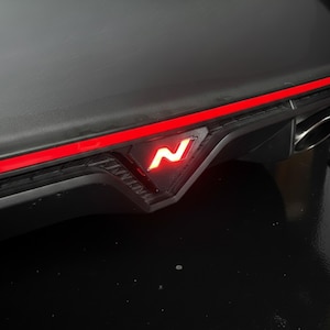 Op de afbeelding: Een matzwarte achterbumperdiffusor voor auto's met een horizontale rode LED-lichtstrip en een centraal driehoekig rood verlicht N-embleem. Het onderdeel bevat een metalen uitlaatopening die zichtbaar is aan de rechterkant van de structuur.