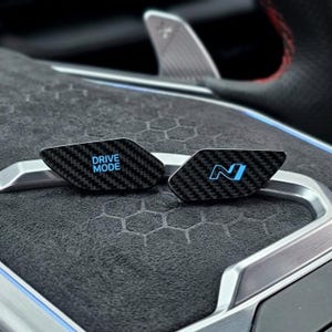May include: Two carbon fibre steering wheel buttons with a black weave pattern. The left button features the text DRIVE MODE in light blue, and the right button displays a light blue N logo. Both buttons are mounted on a dark grey textured surface.