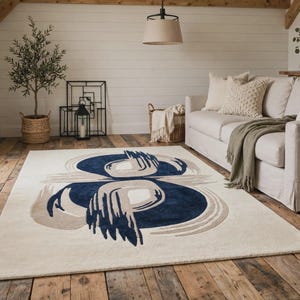 May include: A rectangular area rug featuring a cream-colored base with a central abstract design composed of deep navy blue and light beige brushstroke-style circular patterns that create a symmetrical, bold focal point on the floor surface.
