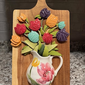May include: A wooden cutting board wall hanging featuring a ceramic pitcher with a painted pink and yellow floral design. The pitcher holds a bouquet of multi-colored tulip-shaped metal cutouts in shades of red, orange, purple, and teal.