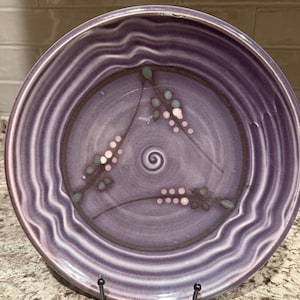 May include: A round ceramic bowl with a purple glaze featuring a wavy, rippled texture on the rim. The center displays a hand-painted spiral design surrounded by three delicate, curved stems adorned with small, light pink and teal circular dots.