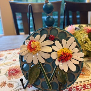 May include: A circular mosaic wall hanging featuring two white daisies with yellow and orange centers, green leaves, and a small red ladybug. The background consists of dark teal circular tiles. It hangs from a jute cord with two teal beads.