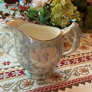 May include: A ceramic creamer pitcher featuring an iridescent finish with a subtle blue and white grid pattern. The pitcher has a gold-trimmed rim, a curved spout, and a sculpted handle with a floral detail at the top, resting on a circular base.