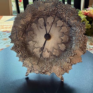 May include: A round metallic table clock with a textured finish featuring a relief pattern of grape clusters and vine leaves around the perimeter. The clock face has a central area with embossed leaf motifs and two thin black hands for timekeeping.