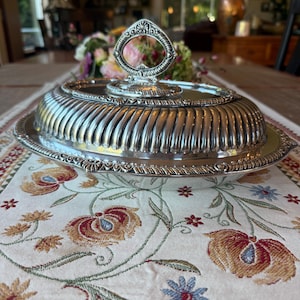May include: An oval silver-plated serving dish with a removable lid featuring a fluted design and a decorative handle. The edges of the dish and the lid are adorned with intricate embossed patterns, providing a polished metallic finish.