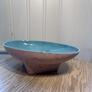 May include: A shallow, oval-shaped bowl with a smooth, light blue interior and a textured, dusty pink exterior featuring a dark, irregular speckled pattern. The bowl rests on a square-shaped base, creating a distinct contrast between the interior and exterior colors.