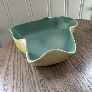 May include: A ceramic bowl with a wavy, organic rim. The interior of the bowl is finished in a smooth, matte teal glaze, while the exterior features a contrasting pale yellow glaze. The bowl has a wide, irregular shape with soft curves.