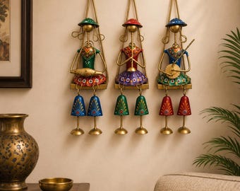 May include: Three wall-mounted metal sculptures depicting figures sitting on swings. Each figure wears a colorful outfit and hat, playing a musical instrument. Below each figure, two bell-shaped ornaments hang from thin metal rods, creating a decorative set.
