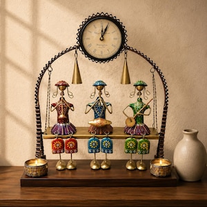 Puede incluir: Un reloj de mesa decorativo de metal con tres figuras de músicos estilizadas sentadas en un columpio. Las figuras están pintadas en rojo, azul y verde y tocan instrumentos. Dos campanas de color latón cuelgan bajo la esfera y dos pequeños portavelas se sitúan sobre la base de madera.