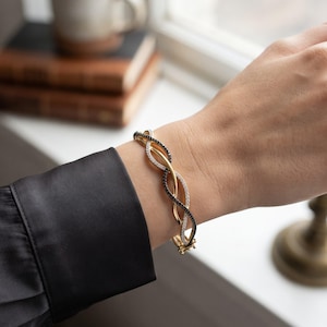 May include: A hinged bangle bracelet featuring a twisted design with three intertwined strands. One strand is polished gold, one is encrusted with clear white stones, and one is encrusted with small black stones, secured with a metal clasp.