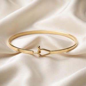 May include: A polished gold-tone bangle bracelet featuring a smooth, circular band that terminates in a unique hook-and-loop style closure. The clasp mechanism consists of a small spherical bead and a teardrop-shaped loop that interlock securely.