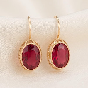 Könnte beinhalten: Ein Paar goldene Ohrhänger mit oval geschliffenen, tiefroten Edelsteinen. Jeder Stein ist in eine dekorative Goldfassung mit Perlenrand und durchbrochener Galerie eingefasst und an einem einfachen, geschwungenen Goldhaken befestigt.