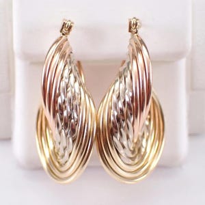 May include: A pair of tri-color gold hoop earrings featuring a layered design. The earrings consist of intertwined yellow, white, and rose gold bands that create a textured, multi-dimensional appearance with a polished finish and secure hinged clasps.