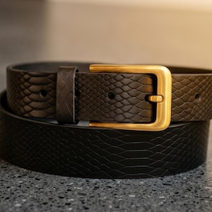 May include: A black belt with a textured snakeskin pattern and a rectangular gold-toned metal buckle. The belt features a single matching loop and multiple punched holes for adjustment, showcasing a consistent dark finish throughout the strap.