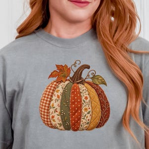 Op de afbeelding: Een grijs t-shirt met korte mouwen voorzien van een centrale afbeelding van een pompoen bestaande uit verschillende stoffen segmenten met patronen in tinten oranje, groen en geel, met een bruine steel en een enkel oranje blad bovenop.