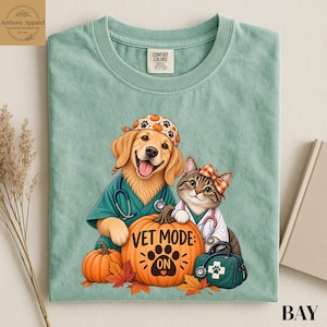 Puede incluir: Una camiseta verde salvia con un golden retriever y un gato atigrado vestidos de veterinarios. El gráfico incluye una gran calabaza naranja con el texto VET MODE: ON, una huella, un estetoscopio y un maletín médico entre hojas de otoño.