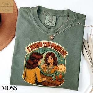 May include: A moss green crewneck t-shirt featuring a graphic print of a person looking into an oval mirror with the text I FOUND THE PROBLEM. The design includes a table with a lamp and a small bottle. The shirt has a Comfort Colors brand label.