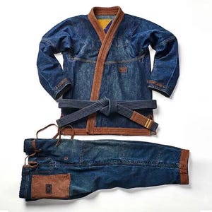 Puede incluir: Un uniforme de artes marciales de mezclilla que consiste en una chaqueta azul oscuro con bordes marrones, mangas largas y un cinturón azul oscuro a juego atado a la cintura. El conjunto incluye pantalones de mezclilla a juego con detalles marrones.