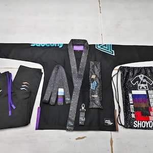 Op de afbeelding: Een zwarte jiu-jitsu gi-set bestaande uit een jas met blauwgroene Saucony-tekst, een gespikkelde kraag, bijpassende broek met paarse trekkoorden, een gespikkelde riem, een kleine sleutelhanger en een zwarte tas met Shoyoroll-tekst.