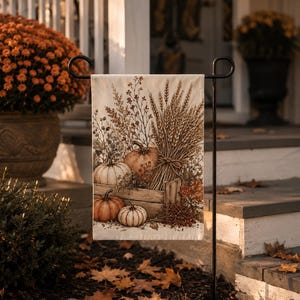 May include: A rectangular garden flag hanging on a black metal stand, featuring a printed illustration of orange and white pumpkins, a wooden crate, and a bundle of wheat stalks with dried foliage in warm autumn tones on a cream-colored background.