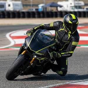 May include: A black and neon yellow motorcycle suit with matching gloves and a full-face helmet, worn while riding a black Yamaha R1 sportbike with neon yellow accents on a paved racetrack with red and white curbing visible in the background.