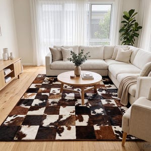 Puede incluir: Una alfombra rectangular con un diseño de parches de cuadrados de piel de vaca en tonos marrón, negro y blanco. La alfombra está colocada sobre un suelo de madera clara en una habitación con un gran sofá seccional crema y una mesa de centro redonda.