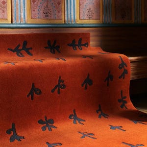 Puede incluir: Una alfombra rectangular con una base de color naranja quemado vibrante y un patrón repetitivo de motivos botánicos abstractos en gris carbón oscuro. La alfombra tiene una textura densa y afelpada, extendida sobre una superficie.
