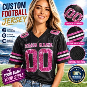 May include: A black football jersey featuring pink glitter lettering that reads TEAM NAME and the number 00. The jersey has short sleeves with pink glitter and white stripes, a V-neckline, and is constructed from a breathable mesh fabric.