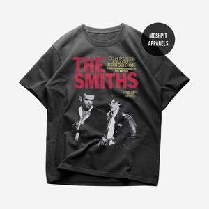 Puede incluir: Una camiseta de manga corta gris carbón con un estampado gráfico de The Smiths. El texto dice THE SMITHS, COMPLETE CHORD SONGBOOK, EVERY SONG RECORDED BY THE SMITHS y COMPLETE WITH LYRICS. Un logotipo circular negro dice MOSHPIT APPARELS.