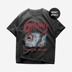 May include: A black short-sleeve t-shirt featuring a graphic print with the red text OBITUARY and CAUSE OF DEATH. The design includes a large eye with a red iris, sharp teeth, and a pile of skulls. A circular black logo reads MOSHPIT APPARELS.
