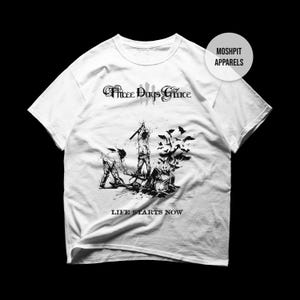 May include: A white short-sleeve t-shirt featuring a black graphic print. The design includes the text Three Days Grace and Life Starts Now, with an illustration of figures and birds. A circular badge reads MOSHPIT APPARELS in the top right corner.