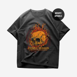 May include: A charcoal gray short-sleeve t-shirt featuring a graphic of a human skull surrounded by orange and red flames. Below the skull, the text reads DISTURBED INSIDE THE FIRE in yellow lettering. A black circular logo reads MOSHPIT APPARELS.