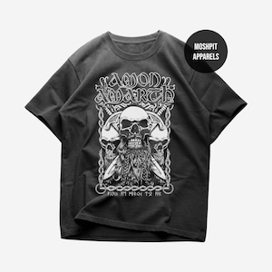May include: A black short-sleeve t-shirt featuring a white graphic print of a bearded skull flanked by two other skulls, swords, and chains. The text reads AMON AMARTH at the top, MOSHPIT APPARELS in a circle, and runic characters at the bottom.