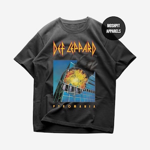 May include: A charcoal gray short-sleeve t-shirt featuring a Def Leppard Pyromania graphic print. The design displays a building with a crosshair target overlay and flames. A circular black badge in the top right corner reads MOSHPIT APPARELS.