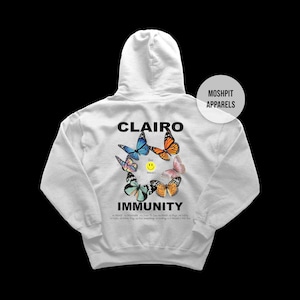 May include: A white hooded sweatshirt featuring a back graphic with the text CLAIRO IMMUNITY, a yellow smiley face, and various colorful butterflies. The text MOSHPIT APPARELS appears in a circular badge. A tracklist is printed at the bottom.