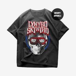 Pode incluir: Uma t-shirt cinzento-carvão de manga curta com o gráfico de um crânio a usar óculos vermelhos com o texto "FREE BIRD" nas lentes. Acima do crânio, o texto "LYNYRD SKYNYRD" aparece em vermelho e branco. Um círculo preto diz "MOSH PIT APPARELS".