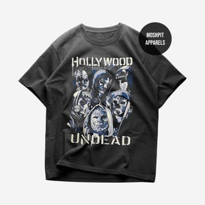 May include: A charcoal grey short-sleeve t-shirt featuring a graphic print of five masked figures in blue and white tones. The text HOLLYWOOD UNDEAD is printed in white above and below the figures. A circular MOSHPIT APPARELS logo is visible.
