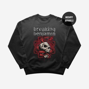 Può includere: Una felpa nera a girocollo con la scritta breaking benjamin in lettere bianche sopra il disegno di un teschio umano avvolto da un serpente rosso. Dietro il teschio c'è un disegno a croce geometrica rossa. È visibile un cerchio nero con testo.