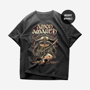 May include: A charcoal gray short-sleeve t-shirt featuring a graphic print of a horned Viking warrior with a beard and an axe, positioned above a skull. The text AMON AMARTH FIRST KILL is printed above the warrior. A circular MOSHPIT APPARELS logo is visible.