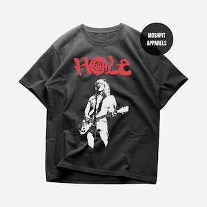 Peut inclure: Un t-shirt noir à manches courtes avec le texte rouge HOLE et un motif en spirale dans la lettre O. Sous le texte se trouve un graphique blanc représentant une personne jouant de la guitare électrique. Un logo noir indique MOSHPIT APPARELS.