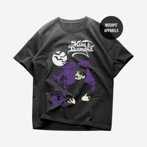 May include: A black short-sleeve t-shirt featuring a graphic print of King Diamond with white face paint and a top hat. The design includes a full moon with bats, a purple silhouette of a house, and the text King Diamond and MOSHPIT APPARELS.