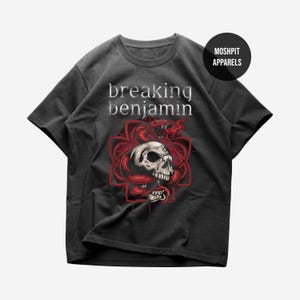 Può includere: Una t-shirt grigio antracite a maniche corte con il testo breaking benjamin in lettere bianche. La grafica include un teschio umano dettagliato avvolto da un serpente rosso, su una croce geometrica rossa. Un cerchio nero contiene il testo MOSHPIT APPARELS.